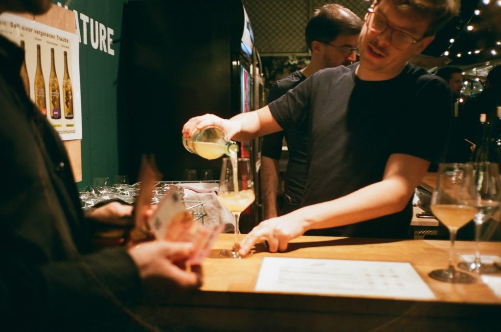 Crop barman in eyeglasses pouring alcoholic drink into glass near anonymous customer with paper money