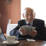 Confident senior businessman holding money in hands while sitting at table near laptop