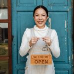 Cheerful ethnic female cafeteria owner in apron demonstrating cardboard signboard while standing near blue shabby door and windows after starting own business and looking at camera
