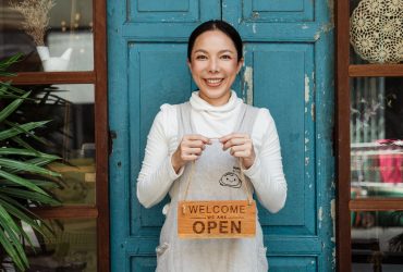 Cheerful ethnic female cafeteria owner in apron demonstrating cardboard signboard while standing near blue shabby door and windows after starting own business and looking at camera