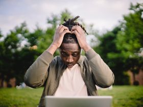 Upset young African American male freelancer clutching head with hands after failure in project while working remotely in park