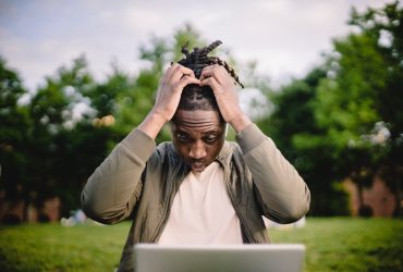 Upset young African American male freelancer clutching head with hands after failure in project while working remotely in park
