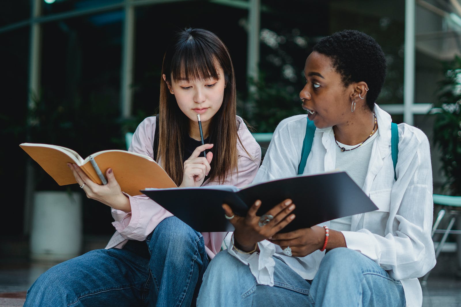 Young black woman talking to Asian girlfriend with copybook while studying together in town